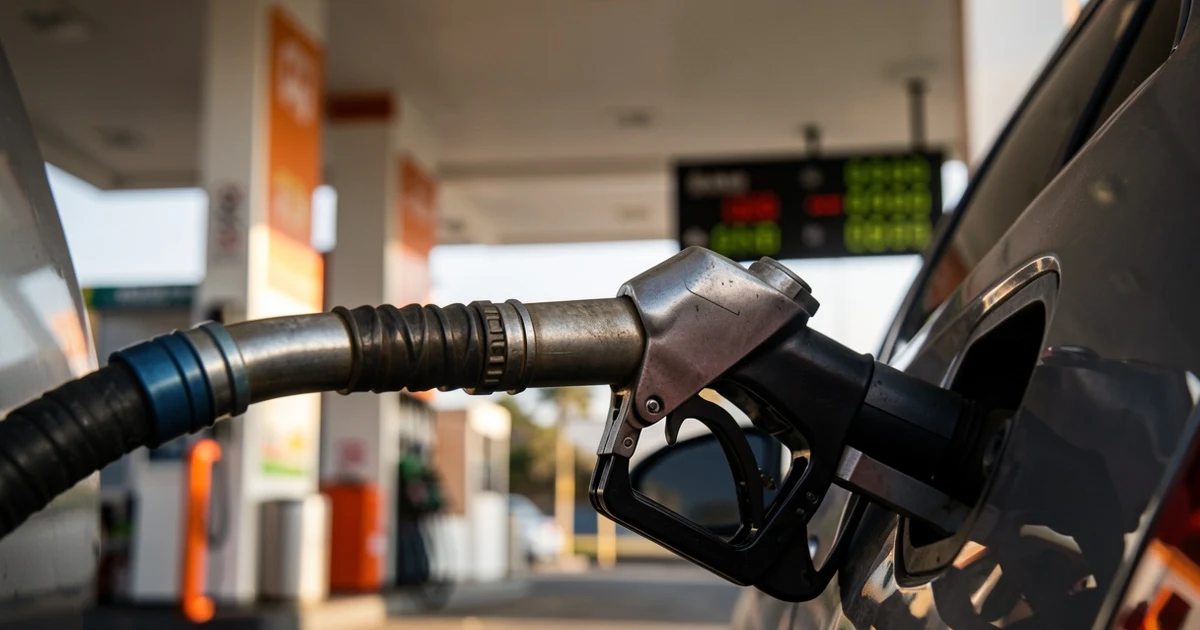 Primer plano de una boquilla de combustible en el depósito de un coche, con fondo desenfocado de gasolinera y cartel de precios
